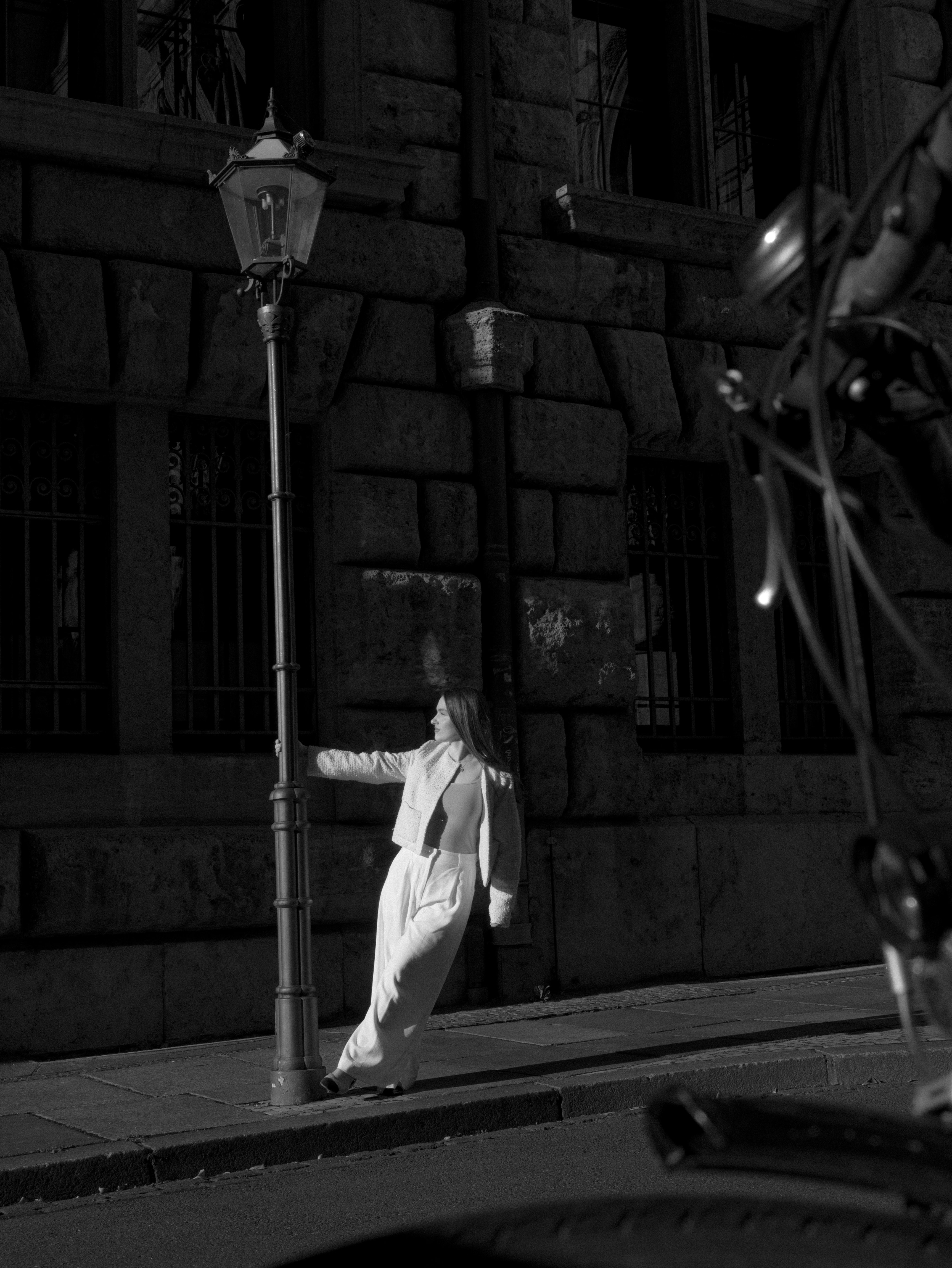 Black-and-white street-style photo of a woman spinning around a lamp post in white trousers and a white jacket