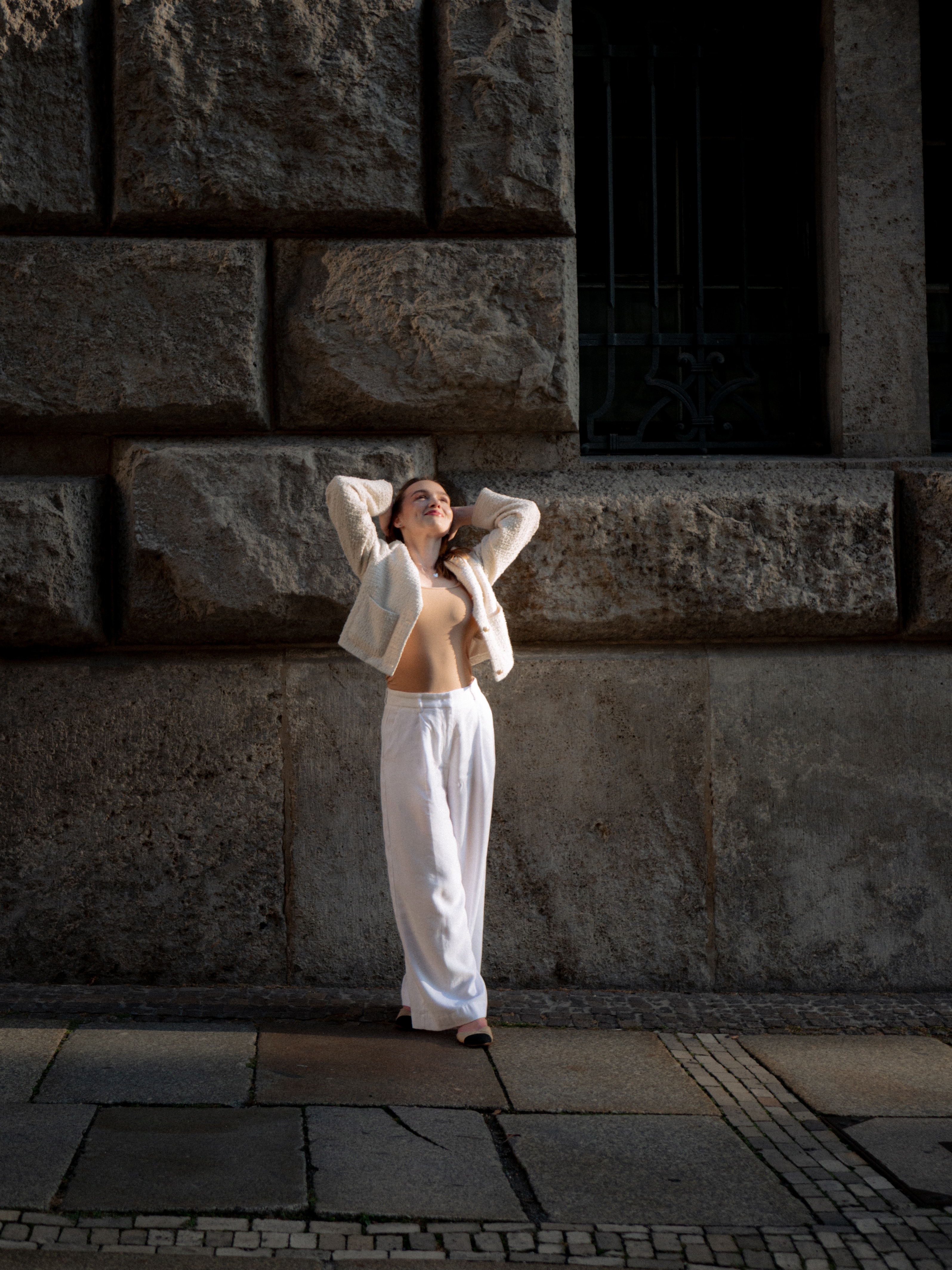 Full-body street portrait of a woman looking upward with her hands on her head, wearing white trousers and a cream jacket against a large brick wall
